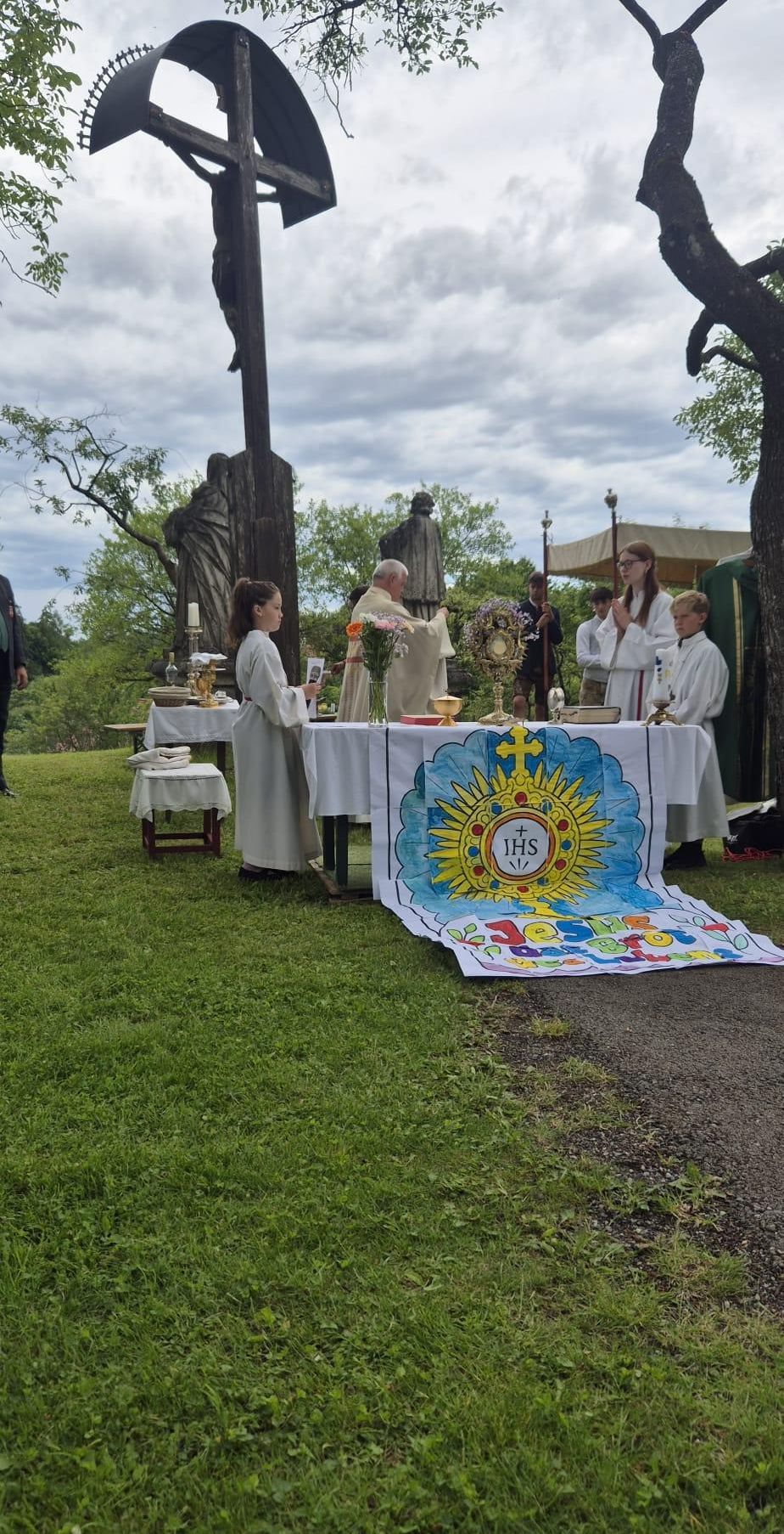Eine religiöse Zeremonie im Freien mit Altären, einem Priester und Altarjungen. Im Hintergrund befinden sich Statuen und Grünpflanzen.