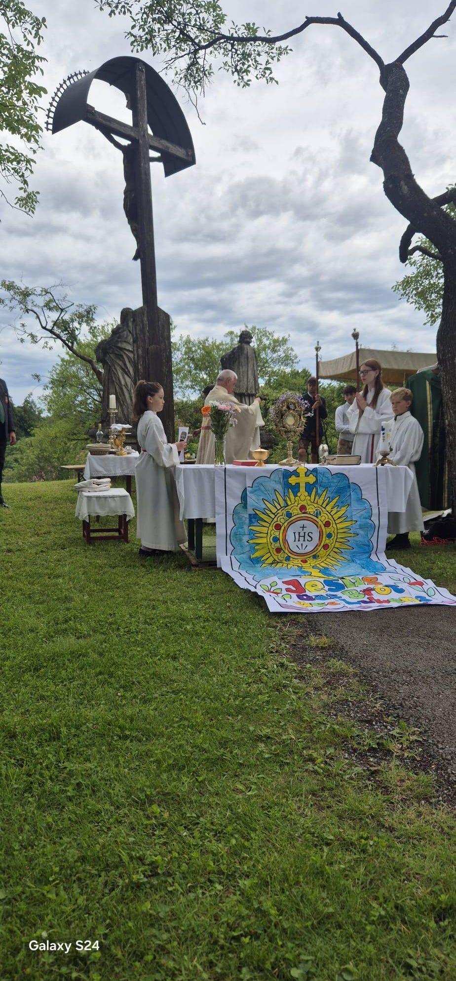 Eine religiöse Zeremonie im Freien mit Altären, einem Priester und Altarjungen. Im Hintergrund befinden sich Statuen und Grünpflanzen.