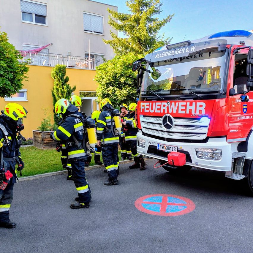 Feuerwehrleute in Schutzkleidung stehen neben einem roten Feuerwehrauto vor einem gelben Gebäude. Bäume und ein Balkon sind im Hintergrund zu sehen.