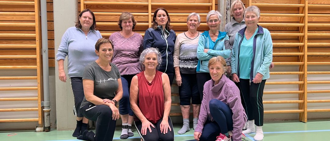 Eine Gruppe von Frauen in Trainingskleidung posiert für ein Foto im Fitnessstudio. Zwei Frauen knien auf Yogamatten, während andere hinter ihnen stehen. Sie alle lächeln und wirken fröhlich.