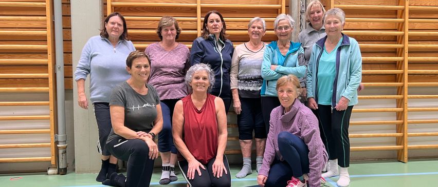 Eine Gruppe von Frauen in Trainingskleidung posiert für ein Foto im Fitnessstudio. Zwei Frauen knien auf Yogamatten, während andere hinter ihnen stehen. Sie alle lächeln und wirken fröhlich.