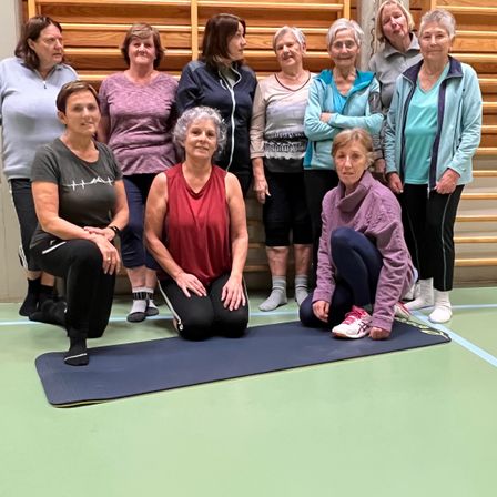 Eine Gruppe älterer Frauen macht Yoga-Posen auf einer blauen Matte in einem Fitnessstudio. Sie tragen Trainingskleidung.