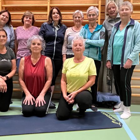 Eine Gruppe älterer Frauen posiert für ein Foto in einem Fitnessstudio. Zwei Frauen knien auf Yogamatten, während andere hinter ihnen stehen.