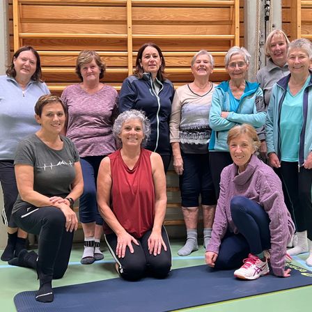 Eine Gruppe älterer Frauen, gekleidet in Sportbekleidung, posiert für ein Foto in einem Fitnessstudio, lächelt und steht auf Yoga-Matten vor einer Holzwand.