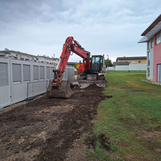 Ein roter Bagger steht auf einer Baustelle neben einem grauen Gebäude und einem Grasfeld. Der Himmel ist bewölkt.