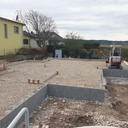 Eine Baustelle mit Kies und Betonblöcken. Ein gelbes Haus und Bäume im Hintergrund. Ein roter Baumaschinenwagen ist geparkt.