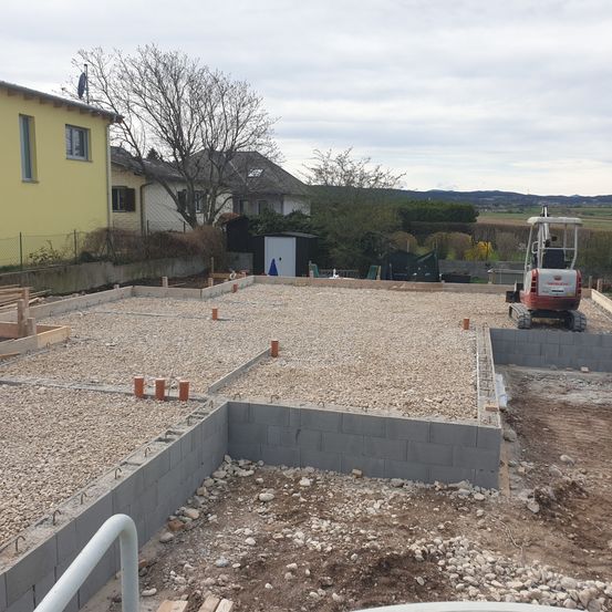 Eine Baustelle mit Kies und Betonblöcken. Ein gelbes Haus und Bäume im Hintergrund. Ein roter Baumaschinenwagen ist geparkt.