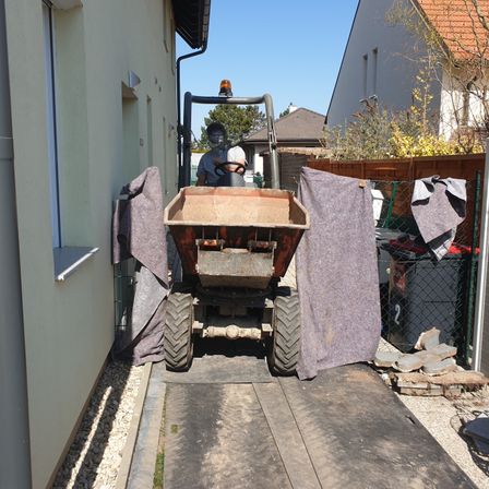 Eine Person fährt mit einem roten Traktor durch eine schmale Gasse, mit einem Gebäude und einem Zaun auf beiden Seiten.