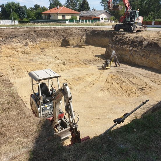 Baustelle mit zwei Baggern und einer Person, die auf einem Erdbereich arbeitet. Ein Haus ist im Hintergrund.