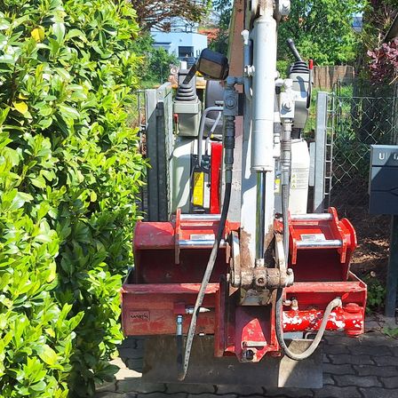 Ein rotes Maschinenfahrzeug steht neben einem Busch und einem Ziegelpflaster. Das Fahrzeug hat einen Metallmast, eine weiße Leitung und einen schwarzen Griff. Es scheint für Wartungsarbeiten verwendet zu werden.