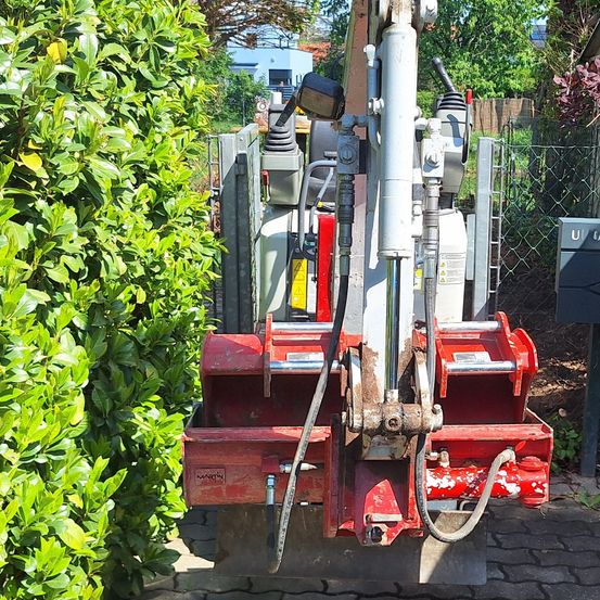 Ein rotes Maschinenfahrzeug steht neben einem Busch und einem Ziegelpflaster. Das Fahrzeug hat einen Metallmast, eine weiße Leitung und einen schwarzen Griff. Es scheint für Wartungsarbeiten verwendet zu werden.