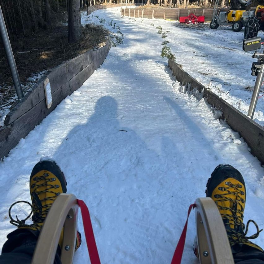 Eine Person benutzt Skier auf einer verschneiten Strecke mit einer hölzernen Barriere an der Seite. Der Schatten des Skifahrers fällt auf den Schnee.