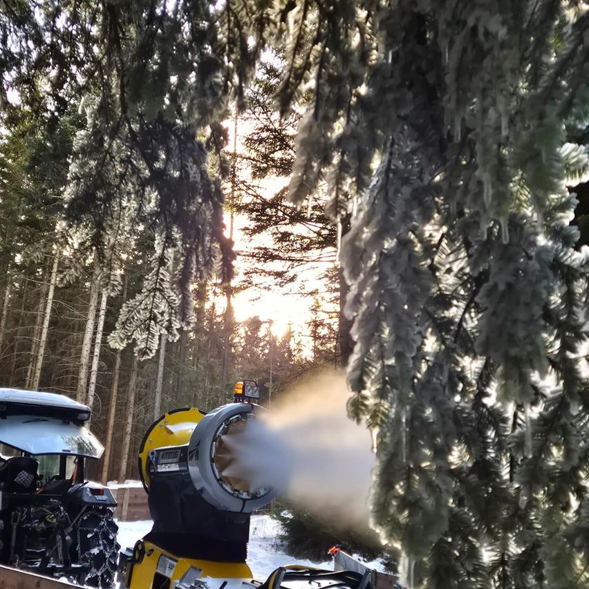 Eine Schneekanone sprüht Schnee in einem verschneiten Wald, mit schneebedeckten Bäumen und einer untergehenden Sonne im Hintergrund.