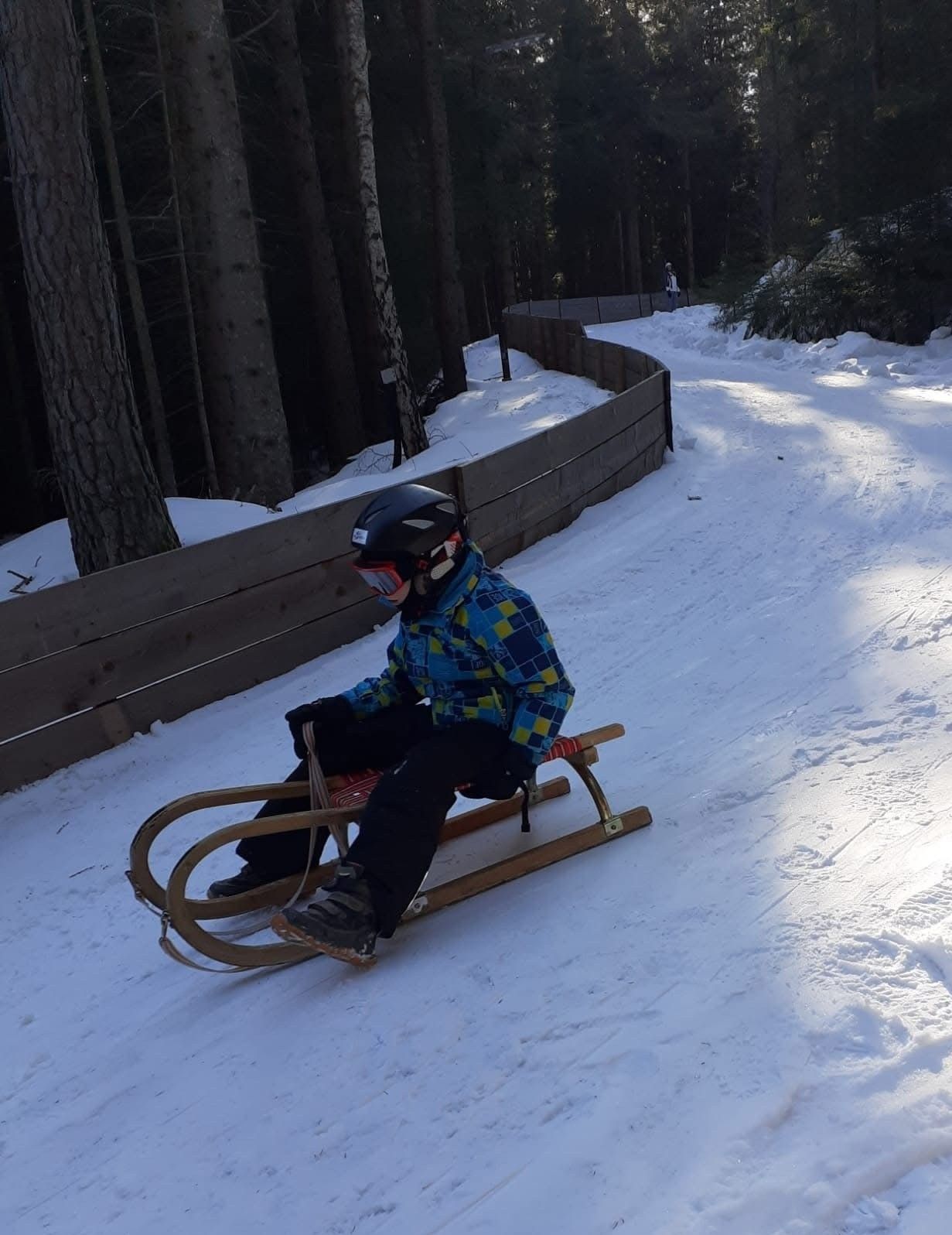 Eine Person in Winterkleidung fährt mit einem hölzernen Schlitten einen verschneiten Hang hinunter, an dessen Seite sich eine Holzbarriere befindet.