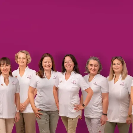 Eine Gruppe von Frauen in weißen Uniformen mit einem Logo auf der Brust posiert für ein Foto vor einem lila Hintergrund.