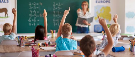 Schüler heben die Hände in einem Klassenzimmer. Eine Lehrerin vor einer Tafel. Das Klassenzimmer hat Schreibtische, Bücher und farbenfrohe Dekorationen.