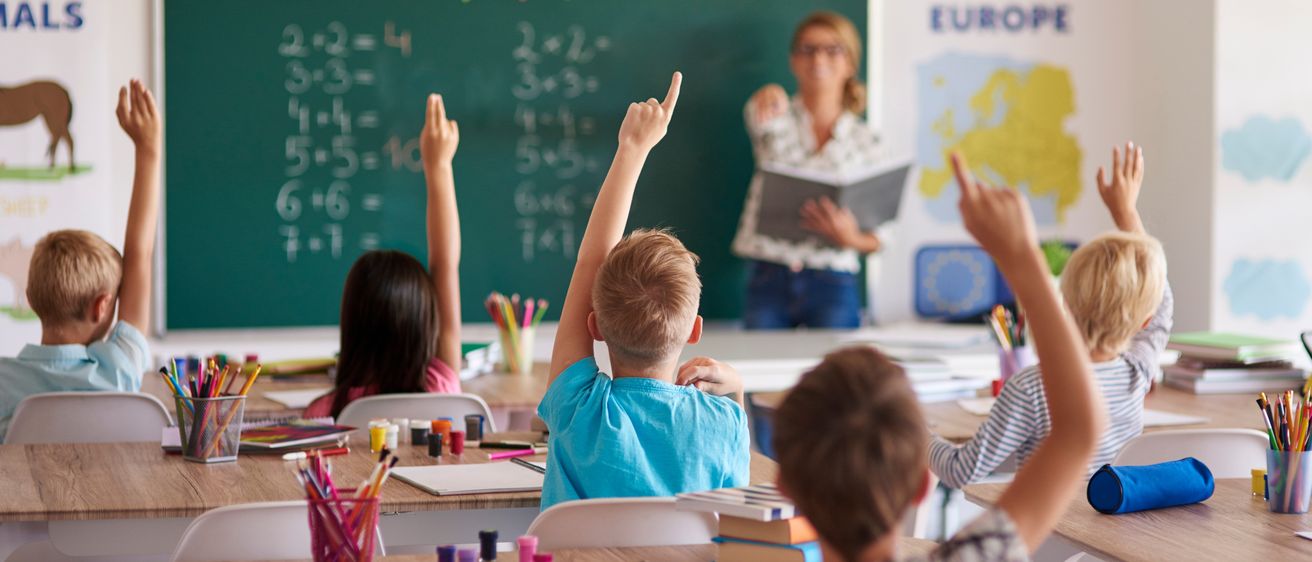 Schüler heben die Hände in einem Klassenzimmer. Eine Lehrerin vor einer Tafel. Das Klassenzimmer hat Schreibtische, Bücher und farbenfrohe Dekorationen.