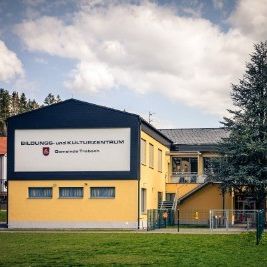 Ein großes gelbes und blaues Gebäude mit einem Schild, das 'Bildungs- und Kulturzentrum' steht in einem Grasbereich mit Bäumen und einem Spielplatz im Vordergrund.