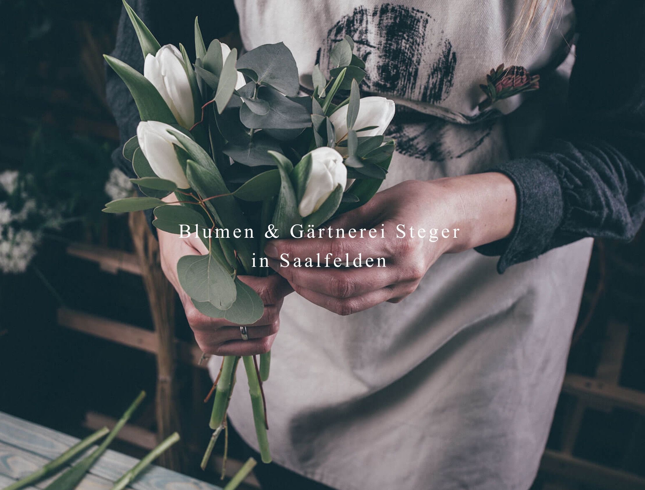 Eine Frau in einer weißen Schürze ordnet einen Strauß weißer Tulpen und grüner Blätter in einer Werkstatt an.
