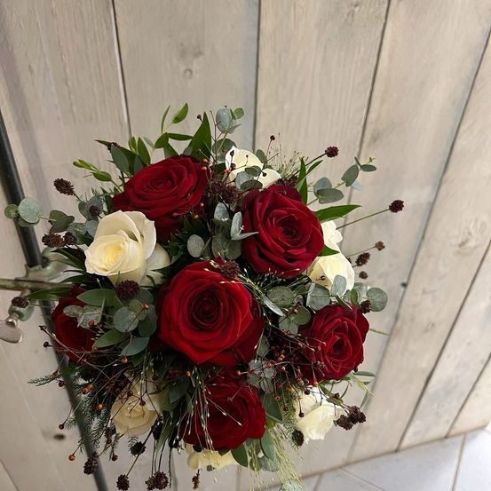 Ein Strauß aus roten und weißen Rosen mit grünen Blättern und kleinen Beeren wird vor einer Holzwand präsentiert.