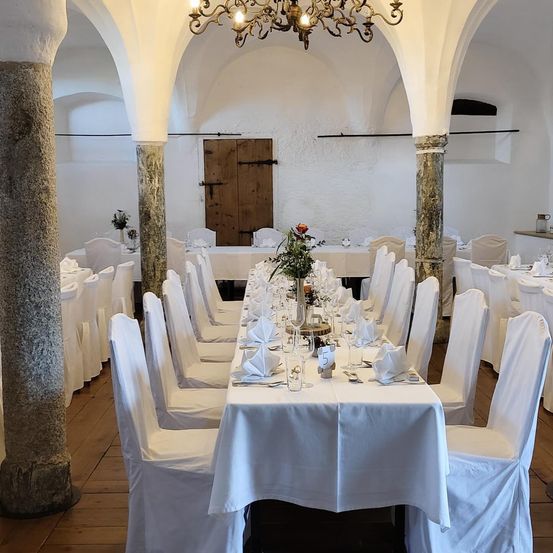 Ein Bankettsaal mit weißen Stühlen und Tischen, die für eine formelle Veranstaltung vorbereitet sind. Der Raum hat Holzböden und weiße Wände mit Steinsäulen. Eine Kronleuchter hängt von der Decke.