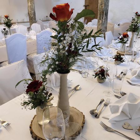 Ein Esstisch mit Blumenarrangements und Besteck, eingerichtet in einem weiß getönten Bankettsaal mit Holzstämmen auf dem Tisch.