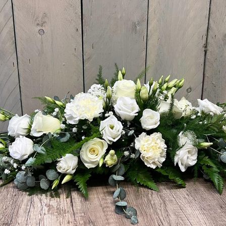 Eine weiße Blumenarrangement mit Rosen und Grünpflanzen liegt auf einem Holztisch vor einer Holzwand.