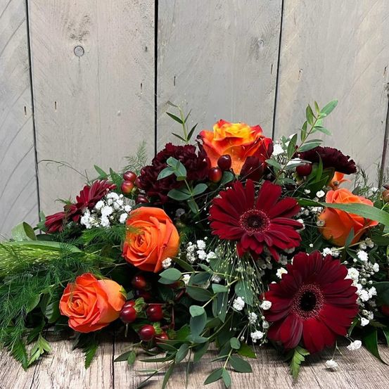 Eine Blumenarrangement aus roten Rosen, burgunderfarbenen Nelken und roten Gänseblümchen ist auf einer hölzernen Oberfläche vor einer Holzwand arrangiert.