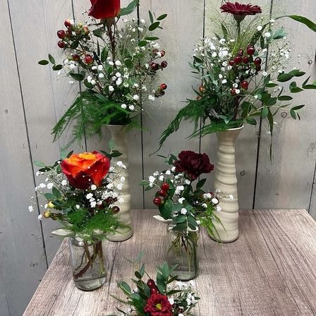 Ein Holztisch hat vier Vasen mit Blumen. Die Vasen sind unterschiedlich groß und farbig. Jede Vase enthält eine einzigartige Blumenanordnung. Im Hintergrund befindet sich eine Holzwand.