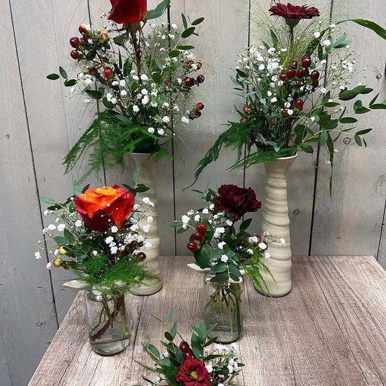 Ein Holztisch hat vier Vasen mit Blumen. Die Vasen sind unterschiedlich groß und farbig. Jede Vase enthält eine einzigartige Blumenanordnung. Im Hintergrund befindet sich eine Holzwand.