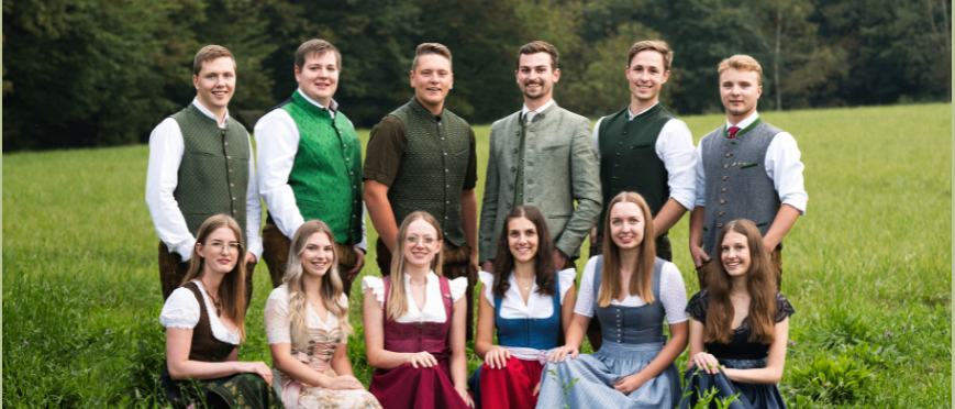 Zwei Bilder zeigen Gruppen von Menschen in traditioneller Kleidung, wahrscheinlich deutscher Tracht. Das erste Bild zeigt Frauen, die in einem Kreis auf einem Grasfeld sitzen. Das zweite Bild zeigt eine Gruppe von Männern und Frauen, die auf einem Feld stehen.