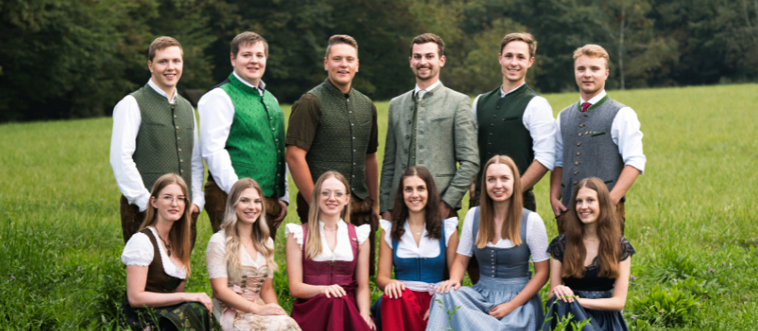 Zwei Bilder zeigen Gruppen von Menschen in traditioneller Kleidung, wahrscheinlich deutscher Tracht. Das erste Bild zeigt Frauen, die in einem Kreis auf einem Grasfeld sitzen. Das zweite Bild zeigt eine Gruppe von Männern und Frauen, die auf einem Feld stehen.