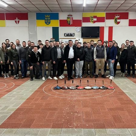 Eine Gruppe von Menschen steht in einem Raum mit einem für ein Spiel markierten Boden. Sie lächeln und posieren für ein Foto. Die Flaggen hängen an der Wand.