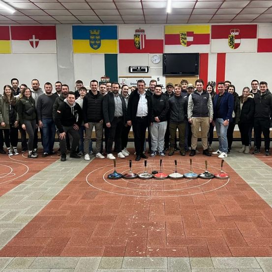 Eine Gruppe von Menschen steht in einem Raum mit einem für ein Spiel markierten Boden. Sie lächeln und posieren für ein Foto. Die Flaggen hängen an der Wand.