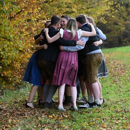 Eine Gruppe von Menschen in traditioneller deutscher Kleidung, einschließlich Lederhosen, schmiegt sich auf einem Grasfeld zusammen, lächelt und lacht. Sie sind von Herbstblättern und Bäumen mit gelbem Laub umgeben.