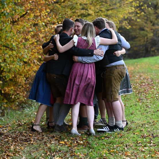 Eine Gruppe von Menschen in traditioneller deutscher Kleidung, einschließlich Lederhosen, schmiegt sich auf einem Grasfeld zusammen, lächelt und lacht. Sie sind von Herbstblättern und Bäumen mit gelbem Laub umgeben.