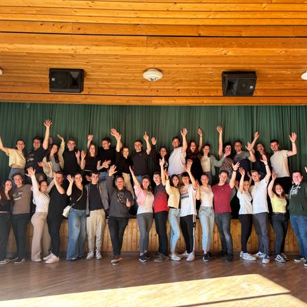 Eine Gruppe von Menschen, die lächeln und winken, posiert für ein Foto in einem Raum mit Holzböden und -wänden. Hinter ihnen hängt ein grüner Vorhang, und zwei Lautsprecher sind an der Wand montiert.