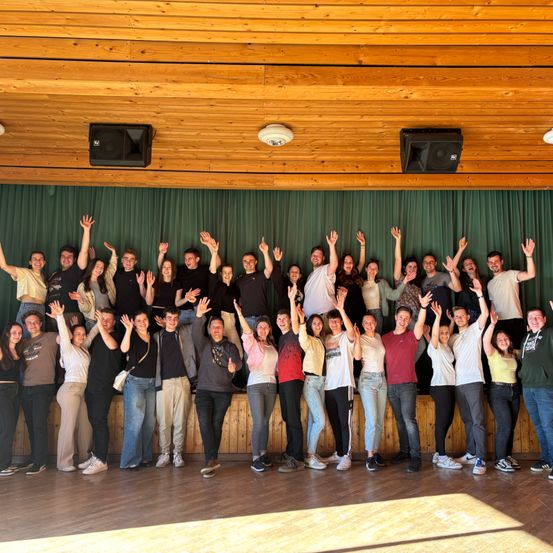 Eine Gruppe von Menschen, die lächeln und winken, posiert für ein Foto in einem Raum mit Holzböden und -wänden. Hinter ihnen hängt ein grüner Vorhang, und zwei Lautsprecher sind an der Wand montiert.