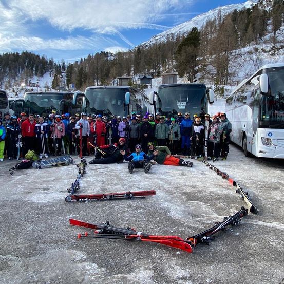 Eine Gruppe von Skifahrern posiert für ein Foto in einem verschneiten Bereich mit Bergen im Hintergrund. Sie bilden ein Herz aus ihren Skiern.