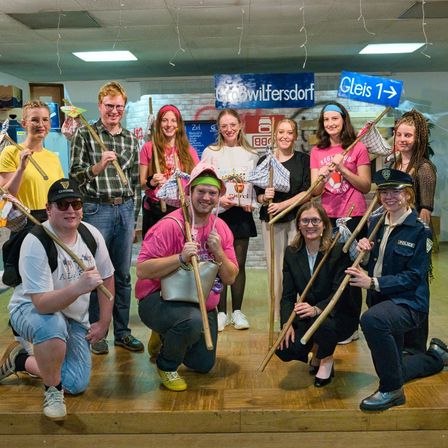 Eine Gruppe von Menschen in Kostümen und mit Requisiten posiert für ein Foto auf einer Bühne. Darunter sind eine Frau in einem rosa T-Shirt, ein Mann in einer blauen Uniform und andere mit Stirnbändern. Hinter ihnen ist ein Banner mit Text.