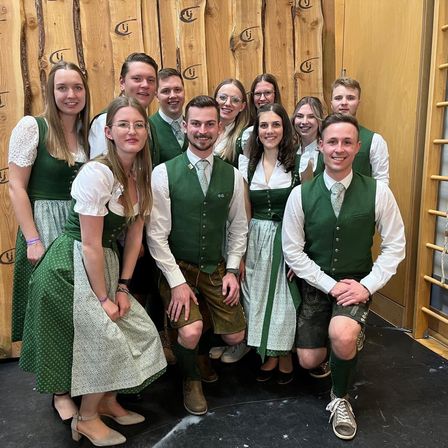 Eine Gruppe von Personen, die in traditioneller grüner Kleidung und Krawatten gekleidet sind, posiert vor einer Holzwand.