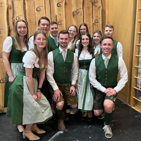 Eine Gruppe von Personen, die in traditioneller grüner Kleidung und Krawatten gekleidet sind, posiert vor einer Holzwand.