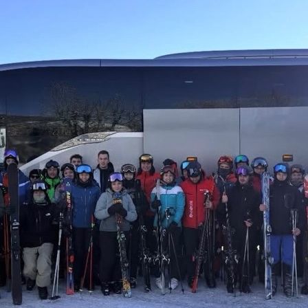 Eine Gruppe von Menschen in Skiausrüstung und Helmen posiert für ein Foto vor einem Bus. Sie halten Skier und Stöcke.