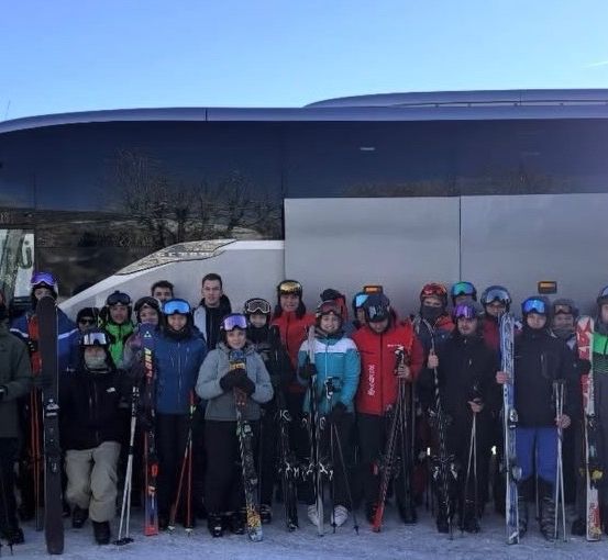 Eine Gruppe von Menschen in Skiausrüstung und Helmen posiert für ein Foto vor einem Bus. Sie halten Skier und Stöcke.