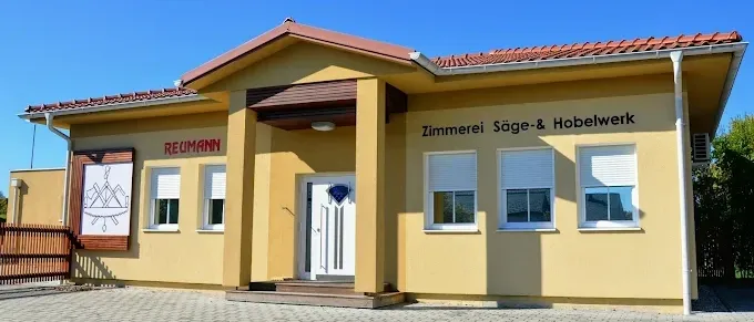 Ein gelbes Gebäude mit rotem Dach und weißen Fensterläden, beschriftet mit 'Zimmerei Sage-Hobelwerk', hat eine weiße Tür mit Diamantmuster.
