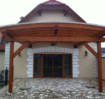 Eine Holzpergola steht vor einem Haus mit einem Ziegeldach, einem steingemusterten Gehweg und einem schwarzen Zaun.