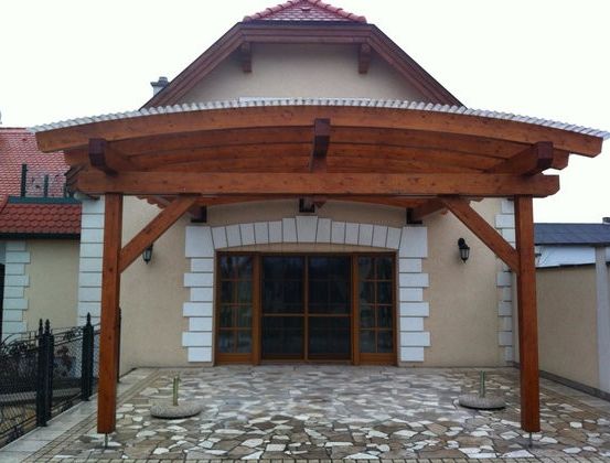 Eine Holzpergola steht vor einem Haus mit einem Ziegeldach, einem steingemusterten Gehweg und einem schwarzen Zaun.