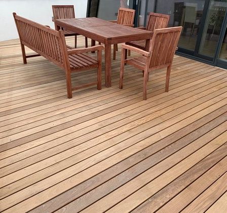 Ein holzfarbener Esstisch ist auf einer Holzterrasse mit Blick auf ein Gebäude und Pflanzen angeordnet.