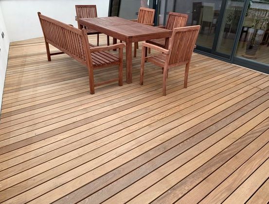Ein holzfarbener Esstisch ist auf einer Holzterrasse mit Blick auf ein Gebäude und Pflanzen angeordnet.