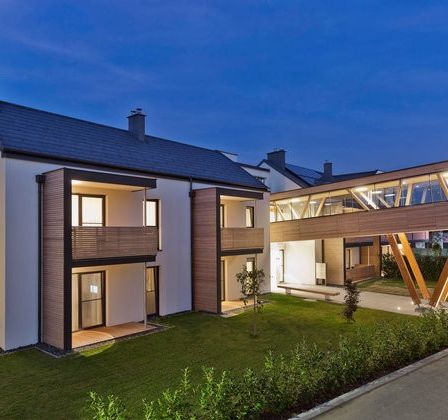 Ein modernes Mehrfamilienhaus mit einer Glasbrücke, die zwei Gebäude bei Einbruch der Dunkelheit verbindet.